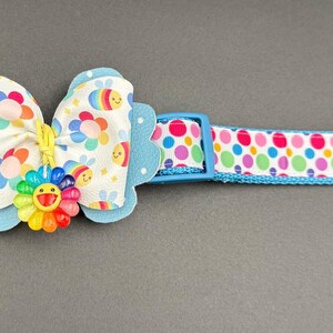 Pode incluir: Uma coleira de cachorro branca e azul com um padrão de bolinhas coloridas. A coleira tem uma fivela azul e um laço combinando com um design de arco-íris e carinha sorridente.