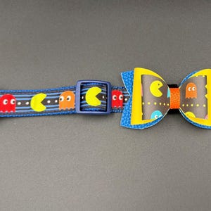 May include: A blue and black dog collar with a Pac-Man pattern and a matching yellow bow tie with a Pac-Man pattern.