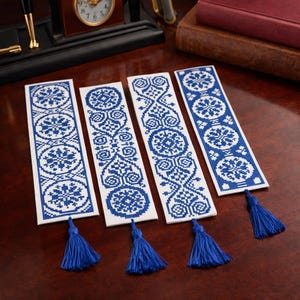 Puede incluir: Cuatro marcadores bordados en blanco y azul con intrincados patrones geométricos y borlas azules. Los marcadores rectangulares presentan un diseño repetitivo de círculos y motivos florales. Están dispuestos sobre una superficie de madera.