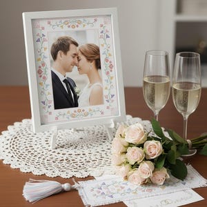 Könnte beinhalten: Ein weißer Fotorahmen mit einem Paar, floralem Rand, steht auf einer Staffelei. Ein Strauß hellrosa Rosen, zwei Sektgläser, eine Deckchen und eine Quaste liegen auf Holz. Hochzeitsthema.