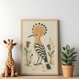 May include: A framed cross-stitch pattern featuring a hoopoe bird with a tan body, black and white striped wings, and a prominent crest, surrounded by purple thistle flowers and tall grasses on a light beige fabric background.