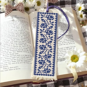 May include: A blue and white embroidered bookmark with a floral design. The bookmark is rectangular with a fringed edge and a purple cord. It is in an open book.