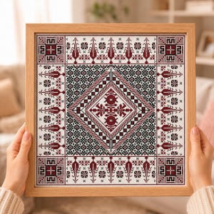 May include: A square cross-stitch embroidery piece framed in light-toned wood. The design features a central diamond motif surrounded by intricate geometric patterns and stylized floral elements in deep red and black thread on a white fabric.