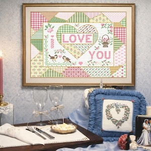 Può includere: Un'opera d'arte a punto croce incorniciata con le parole "LOVE YOU" in rosa, circondata da un design patchwork. Una candela accesa, calici e un vassoio con un pasto sono allestiti per un'occasione romantica. Sono presenti anche un cuscino decorativo e una foto.