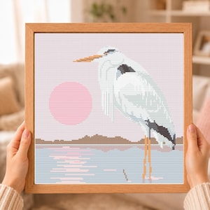 May include: A framed cross-stitch embroidery featuring a white heron standing in shallow water. The scene includes a large pink circular sun, a brown horizon line, and a reflection of the sun on the water, all rendered in a pixelated stitch style.