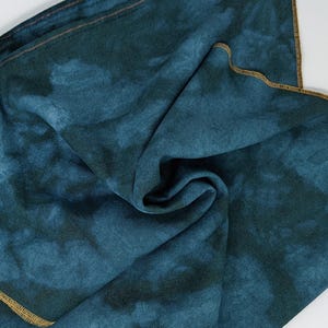 May include: A teal blue fabric with a tie-dye pattern and a gold trim around the edges. The fabric is folded in half and the edges are visible.