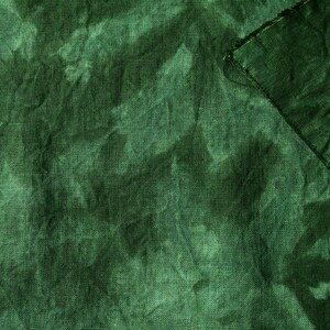 May include: A close-up shot of a textured, dark green fabric. The fabric has a slightly wrinkled appearance, with variations in tone creating a mottled effect. A corner of the fabric is folded over, revealing a raw edge.