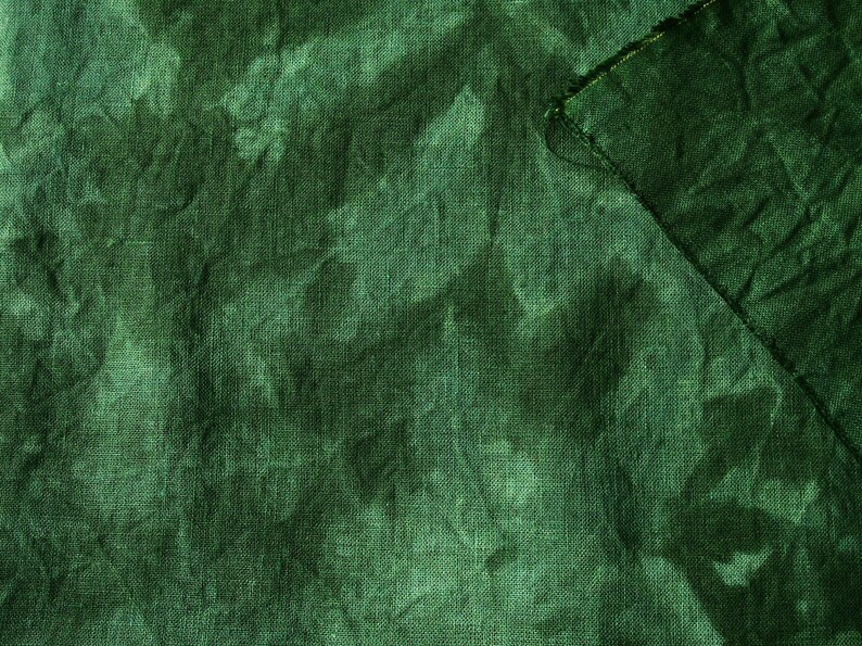 May include: A close-up shot of a textured, dark green fabric. The fabric has a slightly wrinkled appearance, with variations in tone creating a mottled effect. A corner of the fabric is folded over, revealing a raw edge.