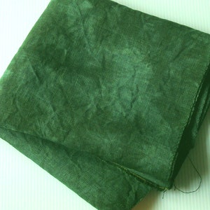 May include: Folded square of dark green linen fabric. The fabric has a textured appearance with visible weave details. The color is a deep, forest green, and the fabric appears to be slightly crumpled, suggesting a natural fiber.