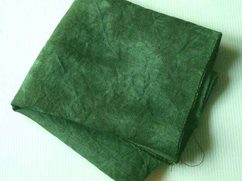 May include: Folded square of dark green linen fabric. The fabric has a textured appearance with visible weave details. The color is a deep, forest green, and the fabric appears to be slightly crumpled, suggesting a natural fiber.
