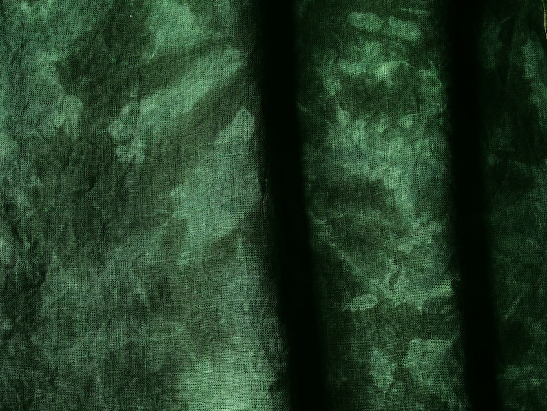 May include: Close-up of a dark green fabric with a tie-dye effect. The fabric has a textured appearance with lighter green patches against a darker green background. Vertical shadows create depth.