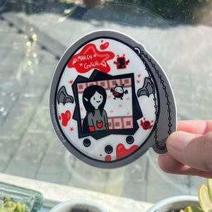 May include: A white and gray circular sticker with a black and white illustration of a character from the animated series "The Owl House". The character is named "Marcy" and is holding a small bat. The sticker has the text "Marcy* Gotchi" in red.