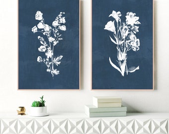 May include: Two framed floral art prints. Each print features a white botanical illustration on a dark blue background. The frames have a light brown finish. The prints are displayed on a white surface with decorative boxes and a small plant.