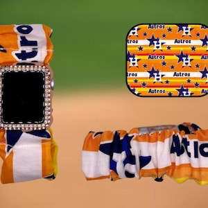 May include: A white and orange Apple Watch band with a blue and white Astros logo. The band is made of fabric and has a scrunchie-like design. The watch face is black and has a diamond-like bezel.