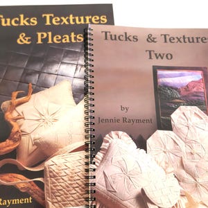 May include: Two spiral-bound instructional books by Jennie Rayment. The left book is titled Tucks Textures & Pleats and features cream-colored fabric designs. The right book is titled Tucks & Textures Two and displays intricate white fabric patterns.