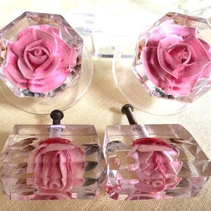 Op de afbeelding: Glazen kastknoppen met roze roosdetails. De knoppen hebben een facetgeslepen, achthoekige vorm en een metalen schroef. De roos is een gedetailleerd, driedimensionaal ontwerp. Decoratieve meubelbeslag.