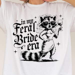 May include: White oversized t-shirt with a black and white illustration of a raccoon wearing a veil and sunglasses, holding a glass of wine. The text "in my feral bride era" is printed on the shirt.