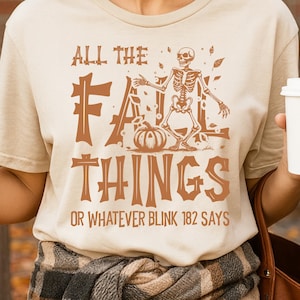 Puede incluir: Camiseta beige con el texto "ALL THE FALL THINGS OR WHATEVER BLINK 182 SAYS" en marrón. El diseño incluye un esqueleto, una calabaza y hojas de otoño. Parte de una colección temática de otoño.