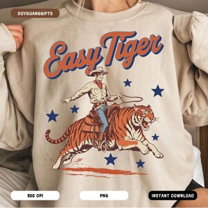 Puede incluir: Una sudadera beige con el texto "Easy Tiger" en una fuente retro. Un vaquero de dibujos animados monta un tigre con un lazo en la mano. La imagen est&aacute; rodeada de estrellas azules.