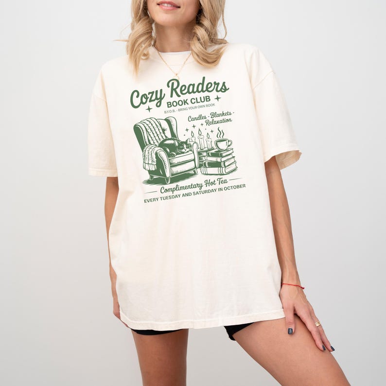 Puede incluir: Camiseta color crema con un gr&aacute;fico verde que dice "Cozy Readers Book Club" con una ilustraci&oacute;n de un sill&oacute;n acogedor, libros, velas y una taza de t&eacute;. El texto tambi&eacute;n incluye "Complimentary Hot Tea" y los d&iacute;as de la semana.