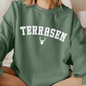 May include: A green sweatshirt with the word "TERRASEN" in white letters and a white deer silhouette.