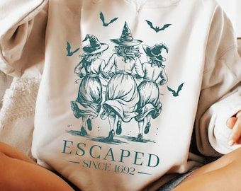 May include: Cream-colored sweatshirt featuring a teal graphic of three witches running, with bats flying above. The text "ESCAPED SINCE 1692" is printed below the illustration. The design is a vintage-style illustration.