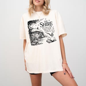 May include: A white t-shirt with a black illustration of a hobbit hole, a pipe, and a book. The text on the shirt reads "The Shire" and "All we have to decide is what to do with the time that is given to us."