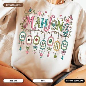 May include: A cream-colored sweatshirt featuring a colorful graphic print that reads "MAHJONG" in stylized lettering. The design includes hanging mahjong tiles, floral vines, butterflies, and small hearts in shades of pink, green, and blue.