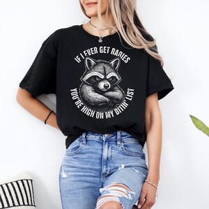 May include: Black t-shirt with a raccoon illustration and the text: "IF I EVER GET RABIES YOU'RE HIGH ON MY BITIN' LIST". The design features a detailed drawing of a raccoon with crossed arms.