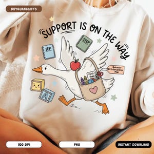 May include: White sweatshirt with a whimsical illustration of a goose carrying a bag filled with school supplies. The goose is flying with the text "SUPPORT IS ON THE WAY" above it. Other elements include an apple, blocks, and notebooks.