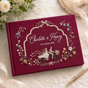 Puede incluir: Un libro cubierto de terciopelo color burdeos profundo con un intrincado bordado blanco de una pareja en una bicicleta tándem, rodeado de motivos florales, fresas y el texto "Charlotte & Henry" con la fecha "18 October 2026" en hilo blanco.