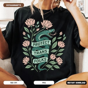Op de afbeelding: Zwart T-shirt met een ontwerp met een groene slang gewikkeld rond een banner met de tekst "PROTECT TRANS FOLKS". Het ontwerp bevat roze lotusbloemen en groene bladeren. Het shirt heeft een ronde hals en korte mouwen.
