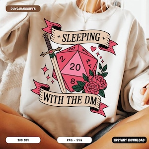 May include: A cream-coloured sweatshirt with a pink D20 die graphic, displaying the number 20. The design includes a sword, roses, and the text "SLEEPING WITH THE DM" on a banner. The sweatshirt has a crew neck.