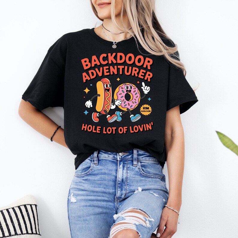 May include: Black t-shirt with the text "BACKDOOR ADVENTURER" in orange and red. A cartoon hot dog and donut are featured in the design, with the phrase "HOLE LOT OF LOVIN'" below. The donut has a "RIM SURVIVOR" badge.