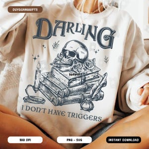 Puede incluir: Una sudadera blanca con un diseño gráfico en blanco y negro que presenta una calavera, flores, libros, una pluma y una vela. El texto "DARLING I DON'T HAVE TRIGGERS" está impreso en la sudadera.