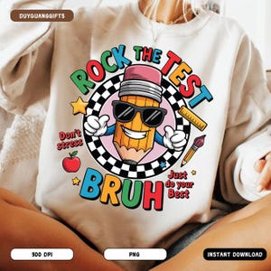 May include: Cream sweatshirt with a vibrant graphic. The design includes a smiling pencil wearing sunglasses, with the words "Rock the Test Bruh." Other elements are an apple, star, ruler, and paintbrush. The sweatshirt is a casual garment.