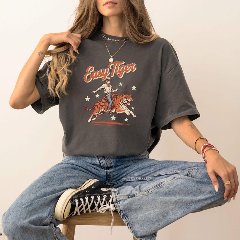 Puede incluir: Una camiseta gris antracita con un gr&aacute;fico vintage de un vaquero montando un tigre. El gr&aacute;fico incluye el texto "Easy Tiger" en una fuente retro.