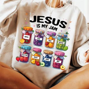 May include: White sweatshirt with the text "Jesus is my jam" printed on the front. The text is surrounded by colorful illustrations of jars filled with fruit and the words "faith", "love", "pray", "justice", "gratitude", "peace", "hope", and "joy".