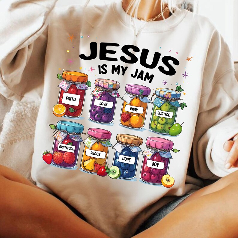 May include: White sweatshirt with the text "Jesus is my jam" printed on the front. The text is surrounded by colorful illustrations of jars filled with fruit and the words "faith", "love", "pray", "justice", "gratitude", "peace", "hope", and "joy".