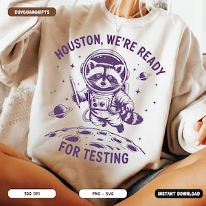 Op de afbeelding: Een wit sweatshirt met een paarse afbeelding van een wasbeer die een astronautenpak draagt en een potlood vasthoudt. De tekst "Houston, We're Ready For Testing" staat op het sweatshirt gedrukt.