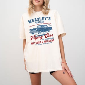 May include: Cream-colored t-shirt featuring a blue vintage car graphic with the text "WEASLEY'S VINTAGE" and "Flying Cars for Adventurous Witches & Wizards". The text is in blue and red, with the location "The Burrow - Devon, England".