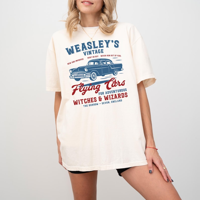 May include: Cream-colored t-shirt featuring a blue vintage car graphic with the text "WEASLEY'S VINTAGE" and "Flying Cars for Adventurous Witches & Wizards". The text is in blue and red, with the location "The Burrow - Devon, England".