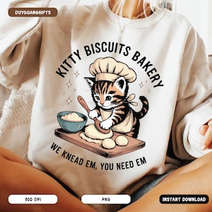 Puede incluir: Sudadera de color crema con un gato de dibujos animados como chef y el texto "Kitty Biscuits Bakery". El gatito está representado amasando masa sobre una tabla de madera. También está presente la frase "We Knead Em, You Need Em".