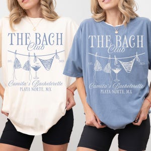May include: Two t-shirts, one cream and one blue, with the text "THE BACH Club" and a bikini and cocktail graphic. The text "Camila's Bachelorette PLAYA NORTE, MX" is also printed on the shirts. The shirts are short-sleeved.