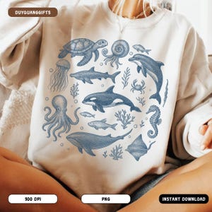 May include: A cream-coloured sweatshirt with a blue illustration of marine life, including a turtle, octopus, dolphin, and whale, in a circular design. The text "DUYQUANGGIFTS" is at the top left.