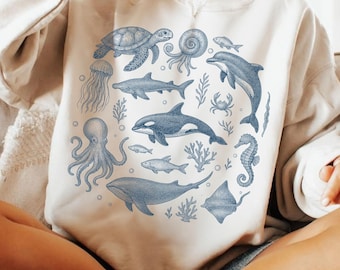 May include: Cream-colored sweatshirt featuring a blue illustration of various sea creatures, including a turtle, octopus, dolphin, and whale, arranged in a circular pattern. The text "DUYQUANGGIFTS" is at the top left.