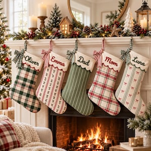 May include: Five fabric Christmas stockings hanging from a white mantel. Each stocking features a scalloped cuff with a name embroidered in red or green: Oliver, Emma, Dad, Mom, and Luna. The stockings have various patterns including plaid, stripes, and small bows, each accented with a green and white gingham ribbon bow.