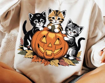 May include: A cream-colored sweatshirt featuring a Halloween-themed graphic. The graphic depicts three kittens of varying colors: black, orange tabby, and gray, posed around a carved orange pumpkin. The pumpkin is surrounded by autumn leaves.