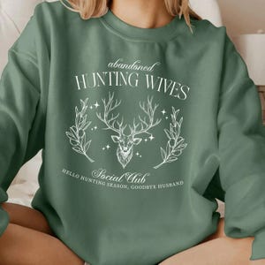 May include: A green sweatshirt with a white graphic design featuring a deer head with antlers and the text "abandoned HUNTING WIVES Social Club HELLO HUNTING SEASON, GOODBYE HUSBAND"