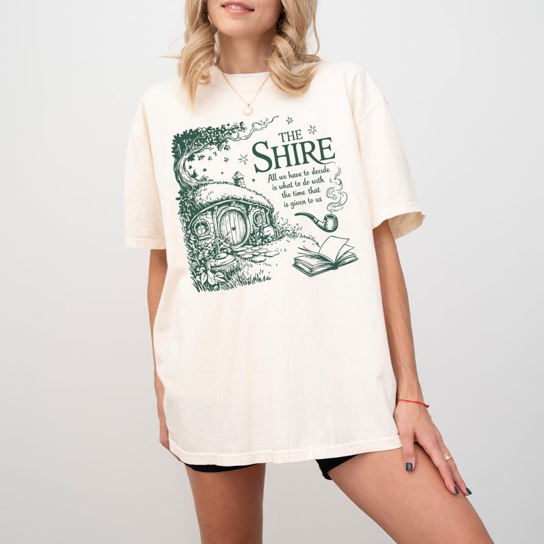 May include: A white t-shirt with a green graphic print of a hobbit house, a pipe, and a book. The text on the graphic reads "THE SHIRE All we have to decide is what to do with the time that is given to us."